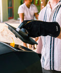 Person using Heavy Duty Heat Resistant Grill Gloves For Outdoor Cooking With Dope Wrist Protection To Stop Burns Ideal For Grilling Barbecues Fireplaces Pizza Ovens And Charcoal Grills to lift grill lid with flames visible.