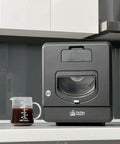 Top Shelf Home Coffee Roaster Machine With Fast Quartz Heating And Ultra Quiet Chaff System For Smooth Daily Roasting In A Sleek Matte Black Cabinet beside a measuring cup.