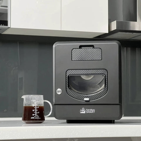 Top Shelf Home Coffee Roaster Machine With Fast Quartz Heating And Ultra Quiet Chaff System For Smooth Daily Roasting In A Sleek Matte Black Cabinet beside a measuring cup.