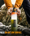 Close-up of hands pressing GeoPress 24 Oz Water Purifier Bottle for hiking and camping against rocks, showing easy fill press to drink design.