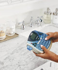 Person using Portable Handheld Label Maker With QWERTY Keyboard And 2 Line Printing on a marble bathroom counter.