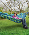 Person relaxing in Extra Long And Wide Double Hammock For Travel Camping Backyard Porch Or Indoor Use With Carrying Pouch Included Blue Green Stripes Ultra Comfy Cotton Weave 142 By 63 Inches Overall And 94 By 63 Inch Bed, hanging between trees on grass.