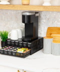 Two Drawer Coffee Pod Organizer With Removable Dividers Fits Up To 72 Pods Or Mix Of Pods And Office Essentials, Metal Mesh Coffee Station Base With Guided Drawer Track For Smooth Opening, Black Finish in kitchen setting with coffee maker and cups on top.