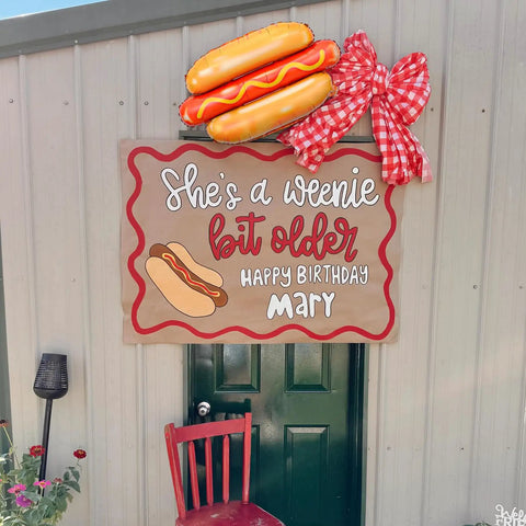 Birthday decor with sign reading 'She's a weenie bit older' adorned with Giant Hot Dog Balloons Set Of 6 28 Inch Food Shape Party Decor.