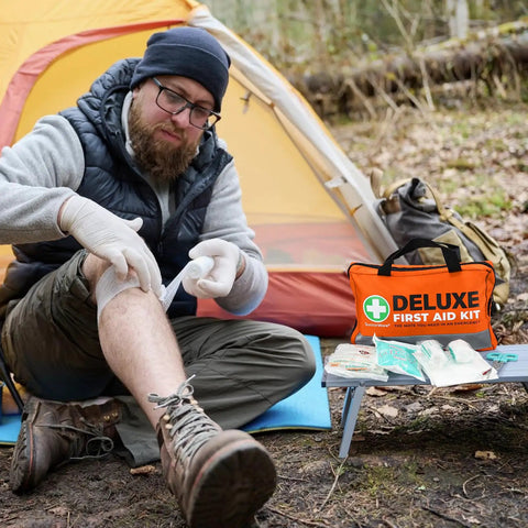 Man using 380 Piece Deluxe First Aid Kit With Bandages Gauze Rolls Instant Cold Packs And Emergency Blanket Compact Orange Kit for outdoor care next to a tent.