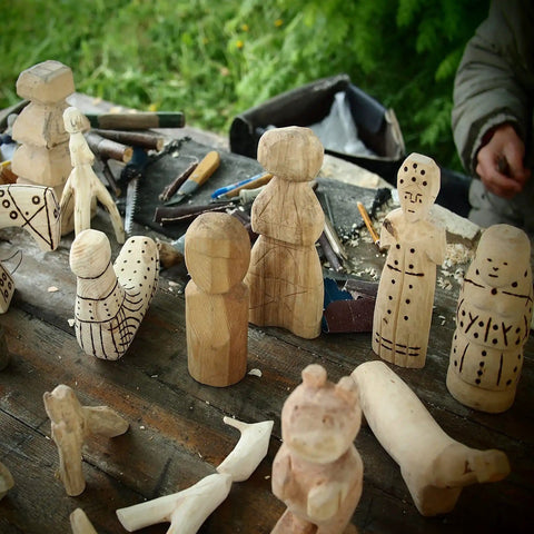 Hand-carved wooden sculptures on a table, showcasing unfinished figurines and carving tools in an outdoor setting.