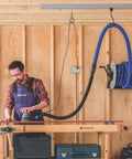 Man working in woodshop with mounted hose reel on wooden wall, tools on workbench, wearing apron.