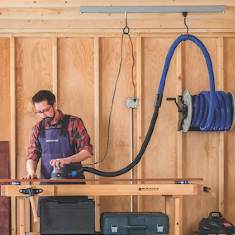 Man working in woodshop with mounted hose reel on wooden wall, tools on workbench, wearing apron.