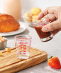 Hand using Two Pack Shot Glass Measuring Cup Set With Thick Base, Multi Unit Liquid Measuring Cup For Espresso And Drinks, Four Measuring Scales Include 1 OZ, 2 TBS, 6 TSP, 30 ML For Precise Pouring