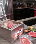 Chamber Vacuum Sealer CM255 For Home And Commercial Kitchens, in a kitchen setting with open lid and steak inside, on a countertop.