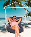 Woman lounging in Cozy Hammock Chair Swing With Wooden Armrests, Sturdy Steel Spreader Bar, Plush Back Pillow on a beach.