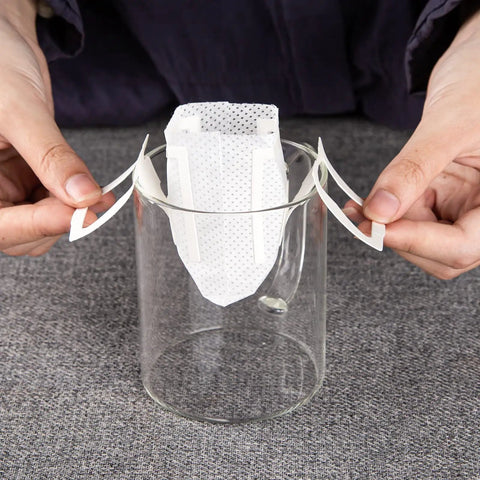 Hands setting up Go To Portable Coffee Filter Bags With Hanging Ear Design on a glass cup for single serve coffee.