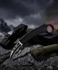 Black karambit knife with carbon fiber handle displayed on rock with strap and paracord, dramatic mountainous background at dusk