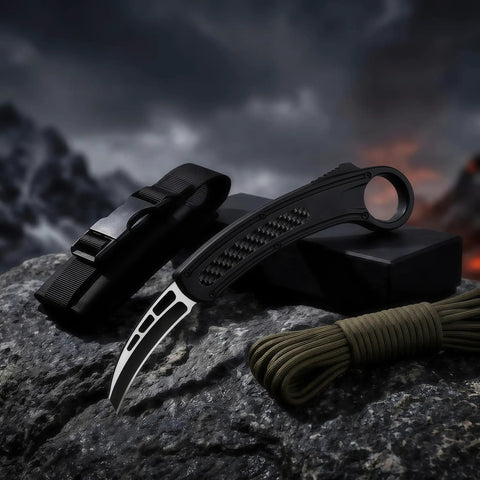 Black karambit knife with carbon fiber handle displayed on rock with strap and paracord, dramatic mountainous background at dusk
