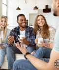 Group of four friends sitting on a sofa enjoying coffee, each holding a mug, in a cozy living room setting with warm lighting.