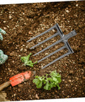 Heavy Duty Steel Farm Fork With Four Prongs for Soil Loosening and Turning, shown on garden soil next to hand trowel