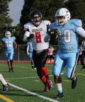 Football player in light blue uniform runs with the ball past an opponent in black and red uniform on a grass field during a game.