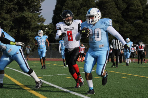 Football player in light blue uniform runs with the ball past an opponent in black and red uniform on a grass field during a game.