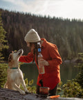 Person in orange jacket holding a portable squeeze water filtration system by a lake with a dog on a fallen tree