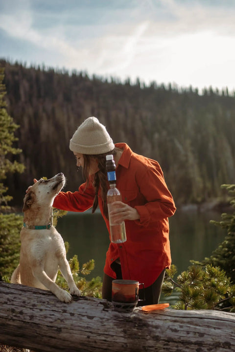 Person in orange jacket holding a portable squeeze water filtration system by a lake with a dog on a fallen tree