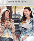 Two women enjoying burgers wrapped in insulated foil wrap sheets, demonstrating greaseproof and leakproof qualities, in a park setting.