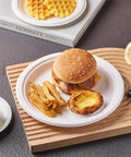 Heavy Duty 9 Inch Disposable Party Plates 150 Pack Eco Friendly Biodegradable Sugarcane Bagasse White Unbleached Dinner Plates For BBQs Picnics And Parties Compostable No Plastic holding a meal with burger, fries, and tart on a textured surface.
