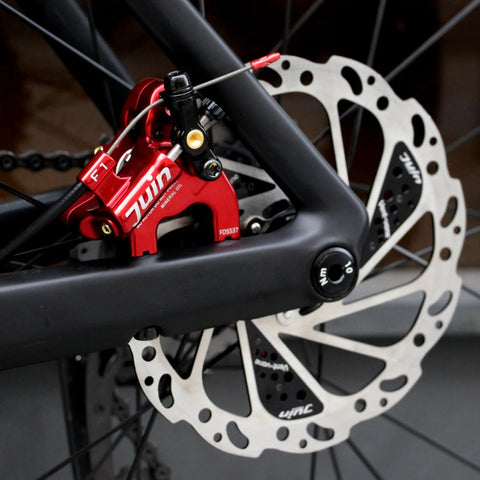 Juin F1 hybrid hydraulic disc brake caliper, red, mounted on a black bicycle frame beside a silver rotor
