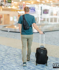 Man at airport with Travel Sling Bag For Daily Use, A Lightweight Crossbody That Is Slim, Compact, And Ready For Commutes Or Travel, Includes Lifetime Lost And Found ID Tag, Water Resistant Black Finish, worn on back, pulling suitcase