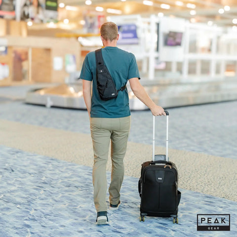 Man at airport with Travel Sling Bag For Daily Use, A Lightweight Crossbody That Is Slim, Compact, And Ready For Commutes Or Travel, Includes Lifetime Lost And Found ID Tag, Water Resistant Black Finish, worn on back, pulling suitcase
