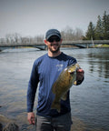 Angler holding a fish by a river with a bridge in the background, wearing a blue shirt and cap.