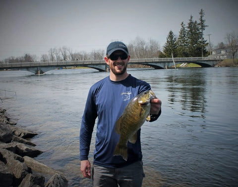 Angler holding a fish by a river with a bridge in the background, wearing a blue shirt and cap.