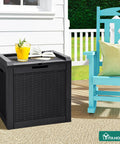 32 Gallon Outdoor Storage Box With Lockable Lid And Side Handles, Waterproof Resin Rattan Deck Box For Patio Cushions, Tools, Pool Accessories, And More, Versatile Side Table Or Storage Ottoman, Black next to a patio chair with a drink on top.