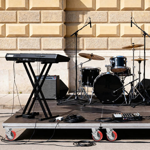 Outdoor music setup with a keyboard on a stand, drum set, and amplifier on a mobile stage with caster wheels visible.