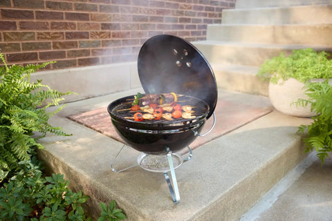 Smokey Joe 14 Inch Portable Charcoal Grill with Tuck N Carry Lid open, outdoor cooking on patio, holding skewers.