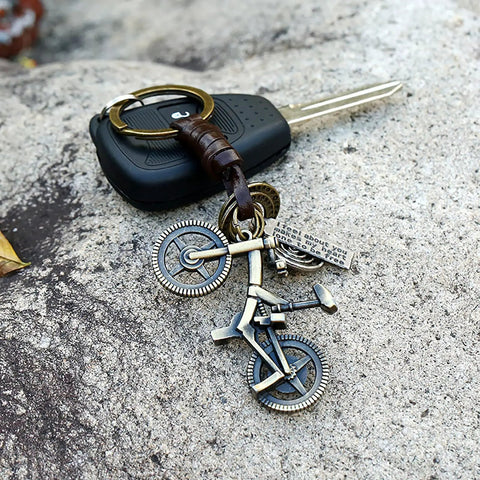 Vintage Leather And Bronze Bicycle Keychain With Metal Wheel And Handlebars Pendant, shown with car key on stone surface.