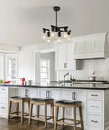Six Light Matte Black Modern Farmhouse Chandelier With Clear Glass Shades hanging above kitchen island with stools.