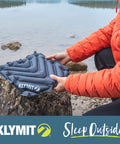 Person holding V Seat Inflatable Cushion For Travel, Camping, Backpacking And Bleachers by a lakeside with rocks.