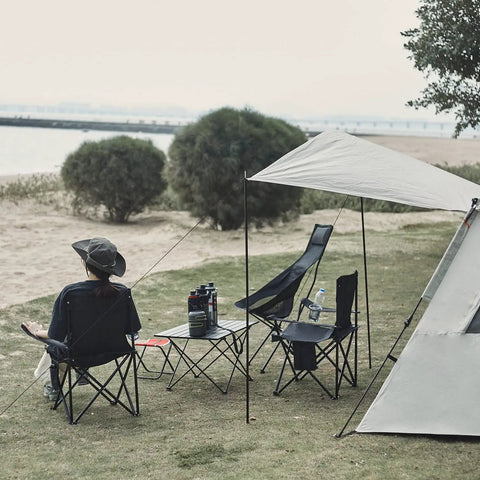 Person sitting on ultra lightweight folding camping chair with carry bag by the beach, surrounded by other camping gear.