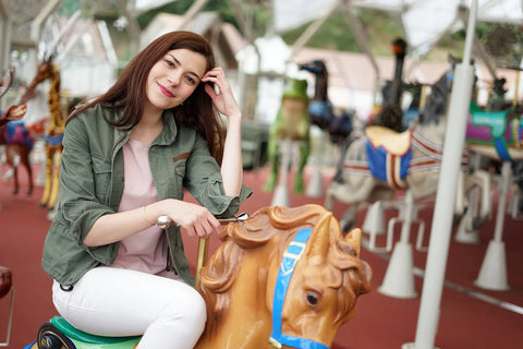Person sitting on a carousel horse, smiling outdoors in a casual outfit with surrounding colorful horses.