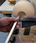 Woodturning tool on lathe shaping a light wood bowl; steel blade, sawdust on the bed.