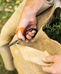 All In One Garden Tool 7-In-1 Pruning Shears in use cutting burlap around plant, showcasing titanium knife and ergonomic handle.