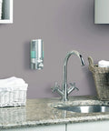Satin Silver One Chamber Wall Mounted Soap And Gel Sanitizer Dispenser in bathroom above countertop with sink and baskets.