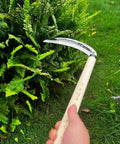 Hand Hoe Sickle Garden Tool For Weeding And Cultivation, sharp blade edge, being used on garden bush, beige handle visible