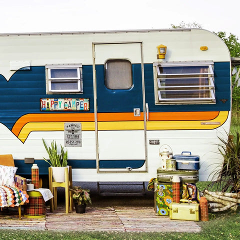 Exterior of a retro camper with a Happy Camper sign. Features colorful decor, plants, and vintage camping gear on a wooden deck.