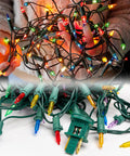 Person holding and displaying brightly lit colorful mini Christmas lights with a plug and connected wires.