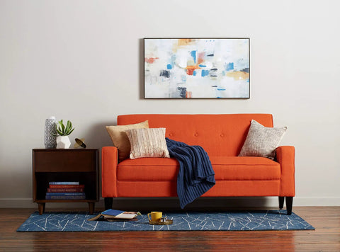 Orange sofa with cushions and a blue throw in a modern living room with abstract wall art above and a wooden side table with books and decor.