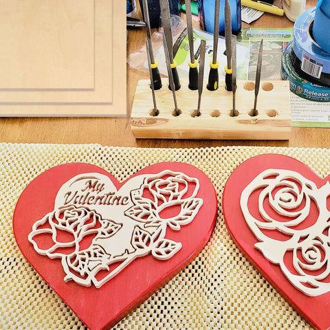 Two red heart-shaped wooden Valentine plaques with white laser-cut roses; drill-bit organizer in the background.