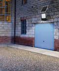 JUDYSSY Solar Flood Lights Led Solar Floodlight mounted on a stone wall, illuminating gravel area near garage.