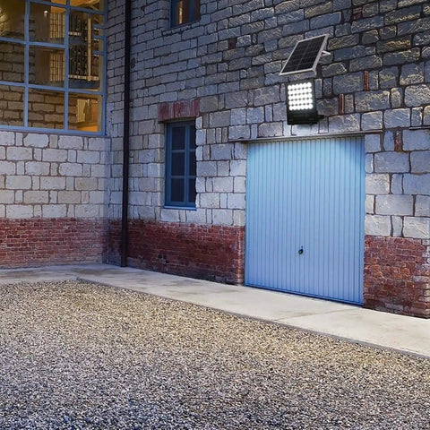 JUDYSSY Solar Flood Lights Led Solar Floodlight mounted on a stone wall, illuminating gravel area near garage.