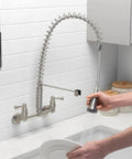 Person using Dual Handle Pro Grade Commercial Style Wall Mount Kitchen Faucet With Pull Down Nozzle And Dual Spray Options In Stainless Steel 1.8 GPM Water Saver For Heavy Duty Kitchens Perfect For Busy Kitchens And Home Chefs, rinsing dishes in a modern kitchen.